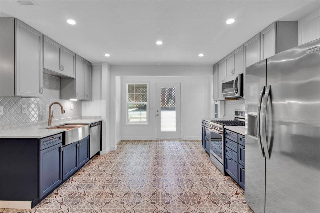 Contemporary kitchen featuring two-tone cabinetry, stainless steel appliances, a farmhouse sink with a bronze-finish faucet, and patterned floor tiles