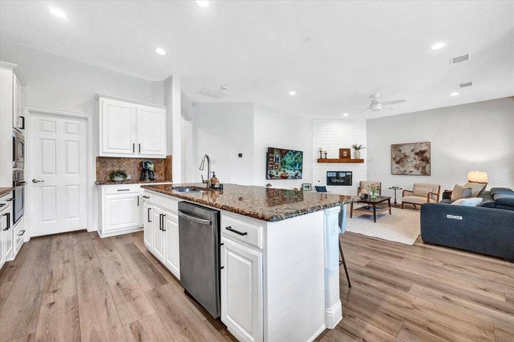 Wood-finish flooring throughout, kitchen island with granite countertop and integrated sink, stainless steel dishwasher, white cabinetry with black hardware, and a living area with a fireplace featuring a shiplap accent wall