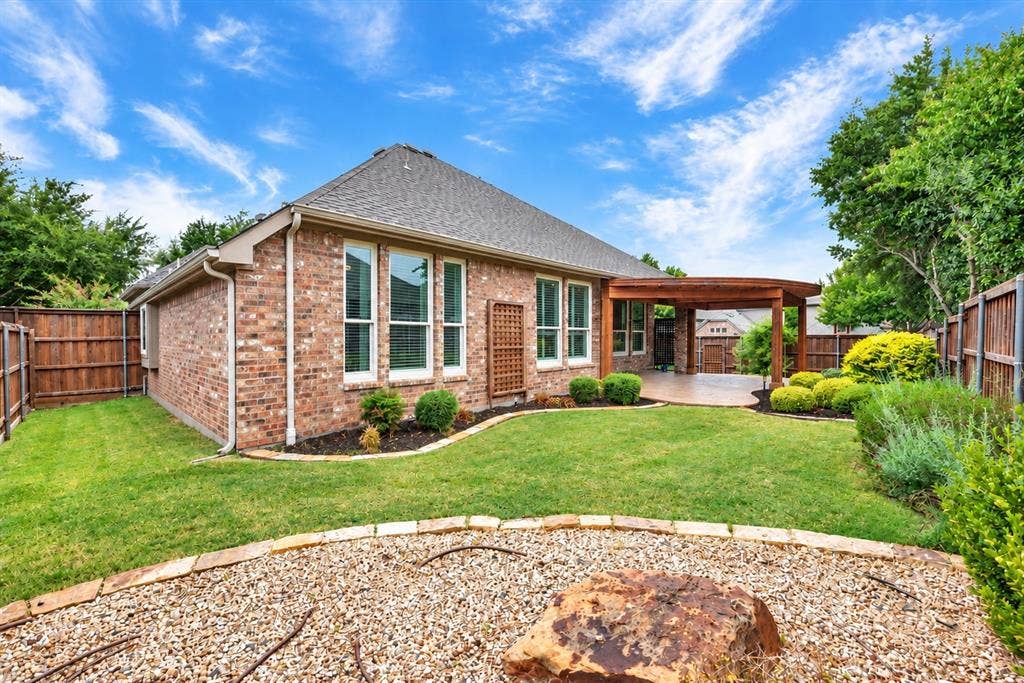 Red brick exterior with white trim, covered patio featuring a wood pergola, expansive lawn, established perimeter landscaping, and a gravel bed with a large stone feature