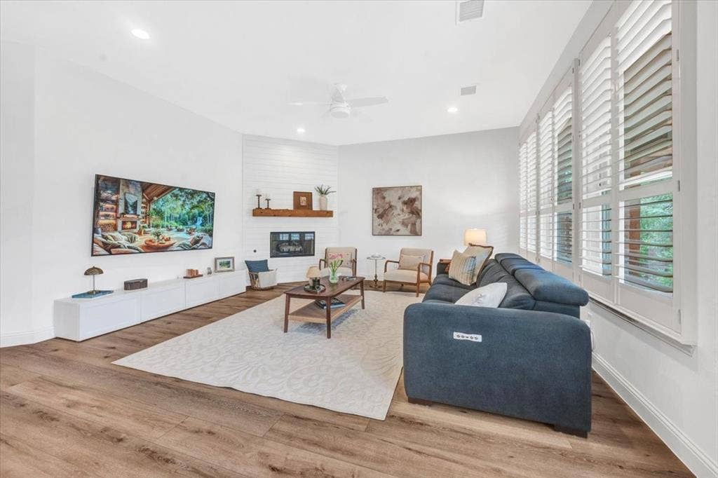 Spacious living area featuring wood-finish flooring, a white shiplap fireplace with a wood mantle, recessed lighting, and multiple windows with plantation shutters