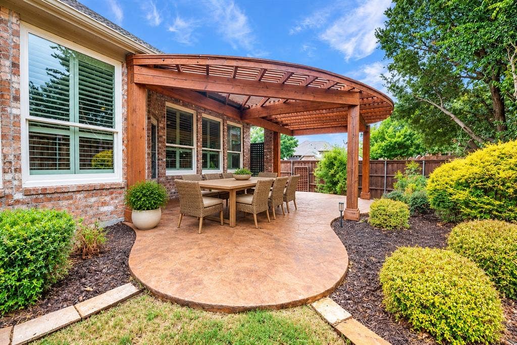 Curved wood pergola over a stamped concrete patio, brick exterior with multiple windows, and mature landscaping