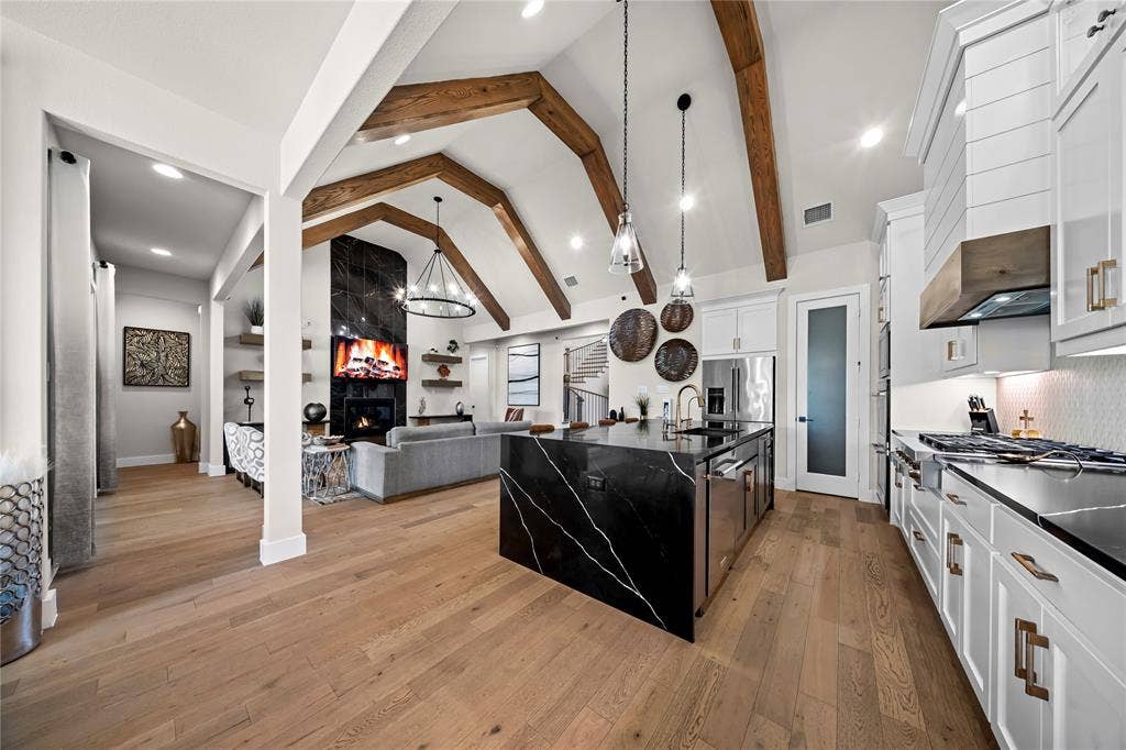 467 Mayfair Lane, Frisco, TX 75033 Vaulted ceilings with exposed wood beams, wood-finish flooring, black stone kitchen island with waterfall edge, white shaker cabinetry, and a black stone fireplace surround