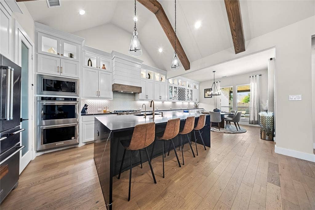 467 Mayfair Lane, Frisco, TX 75033 Vaulted ceiling with exposed wood beams, wood-finish flooring, custom white cabinetry, stainless steel double oven, and a large kitchen island with seating