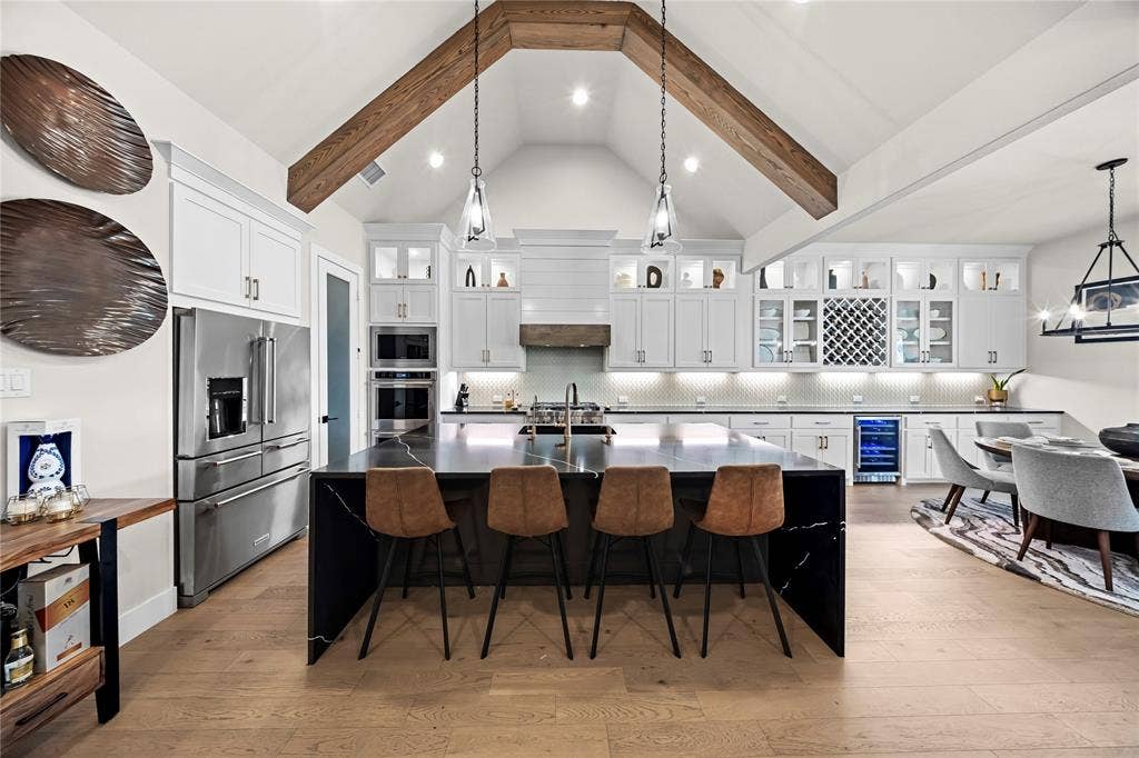 467 Mayfair Lane, Frisco, TX 75033 Vaulted ceiling with exposed wood beams, large kitchen island with waterfall counter, built-in wine storage, stainless steel appliances, and wood-finish flooring