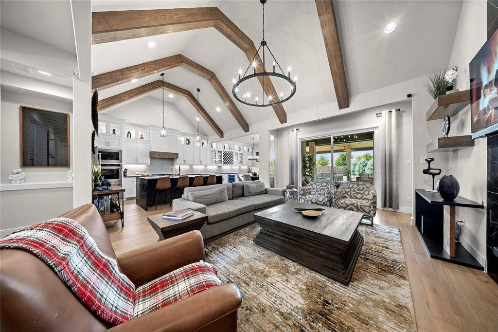 467 Mayfair Lane, Frisco, TX 75033 Vaulted ceilings with exposed wood beams, wood-finish flooring, a kitchen with white cabinetry and built-in appliances, and a sliding glass door to an outdoor living area