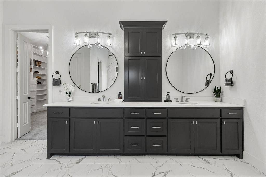 1561 Saint Peter, Prosper, TX 75078 Luxurious dual-vanity bathroom featuring two circular framed mirrors with integrated lighting, extensive black cabinetry with multiple drawers and storage, polished stone flooring with linear patterning, chrome faucetry, and a tall black linen cabinet