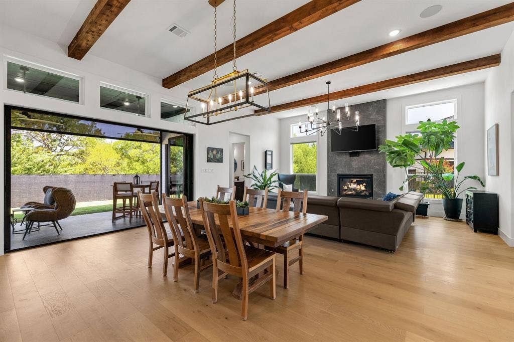 1561 Saint Peter, Prosper, TX 75078 Vaulted ceiling with exposed wood beams, wide plank wood-finish flooring, full-height accordion glass doors, modern fireplace, and a stone-clad fireplace surround