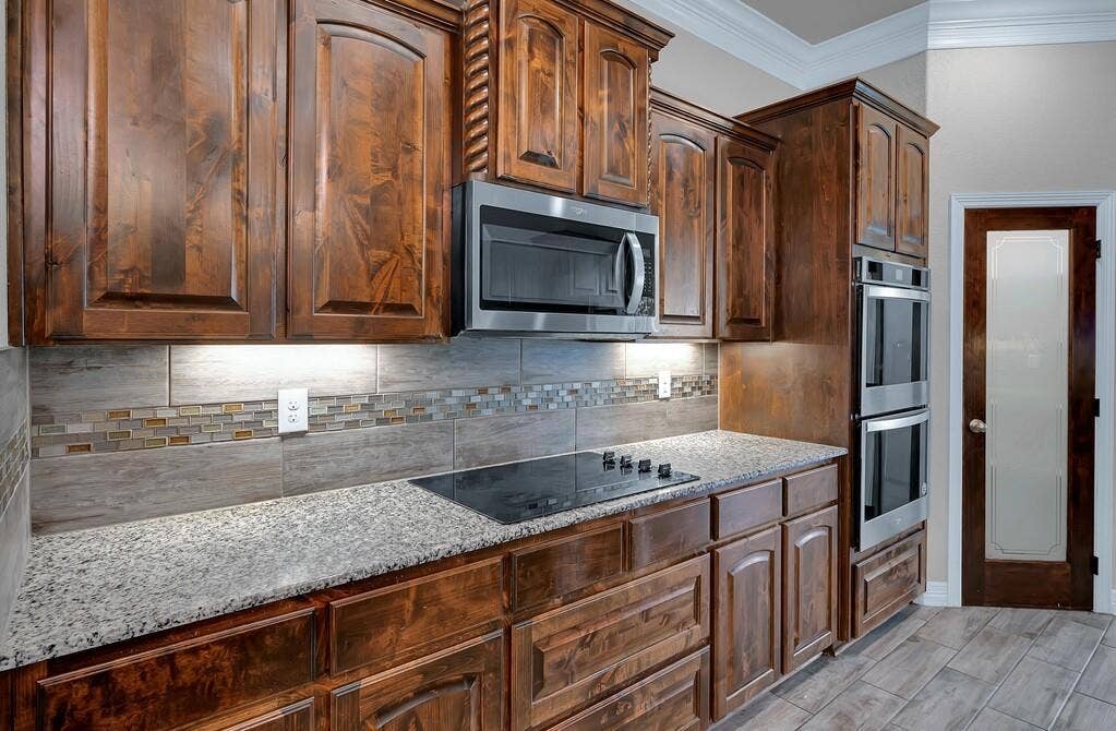 279 Sportsman Drive, Lorena, TX 76655 Warm wood cabinetry with crown molding, stainless steel microwave, integrated cooktop, stone countertops, and tile backsplash with decorative mosaic accent