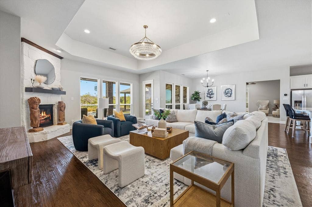 Expansive living area featuring wood-finish flooring, a white brick fireplace with a dark mantel, a tray ceiling, and large windows