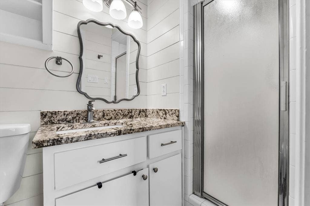 1214 Baylor Avenue, #109, Waco, TX 76706 White vanity with a stone countertop and integrated sink, framed mirror, wall-mounted vanity lighting, and a glass-enclosed shower