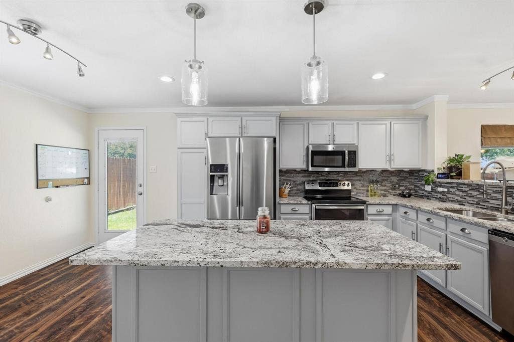 800 Poncho Lane, Fort Worth, TX 76052 Chef's kitchen featuring custom gray cabinetry, granite countertops, a large waterfall-edge island with seating, stainless steel appliances including a French-door refrigerator and gas range, mosaic tile backsplash, pendant lighting, and hardwood flooring throughout the open layout with direct access to the backyard