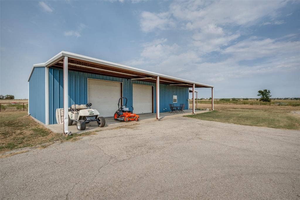 8400 Holder Road, Northlake, TX 76247 Barndo -Blue metal building with two garage doors, an extended covered porch, a concrete driveway, and an expansive open landscape