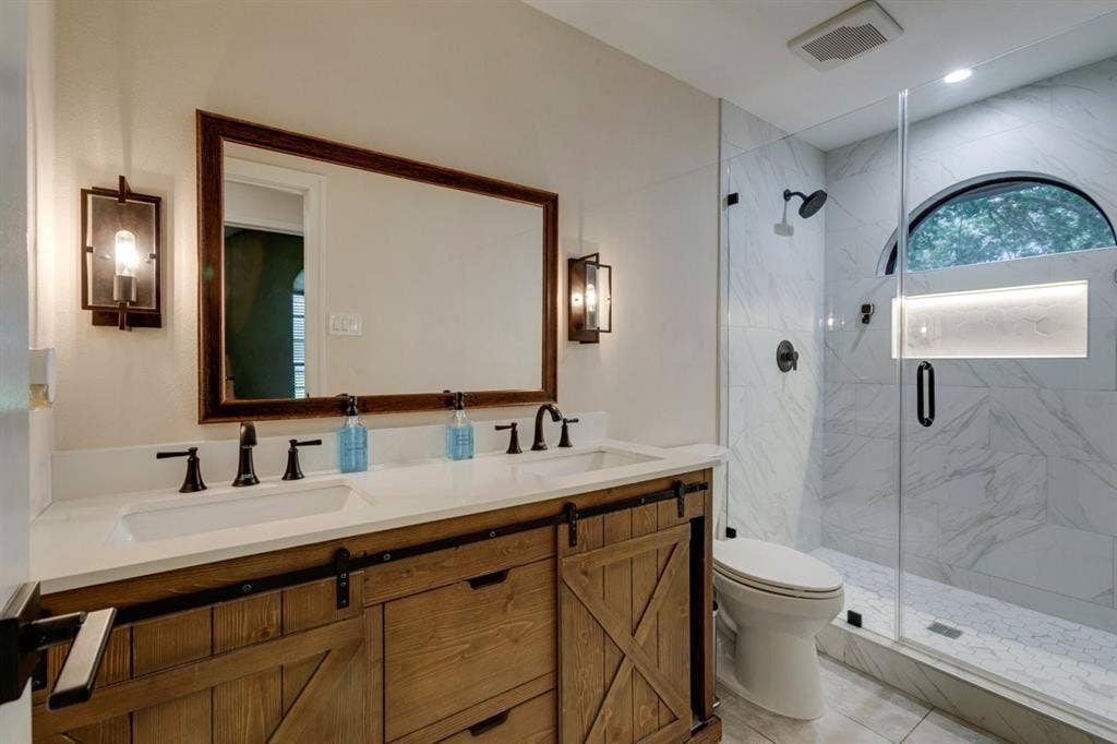 1408 Quapaw Trail, Mesquite, TX 75149 Dual vanity with a white countertop and matte black fixtures, barn door-style cabinet, walk-in shower with glass enclosure, arched window, and marble-look wall tile