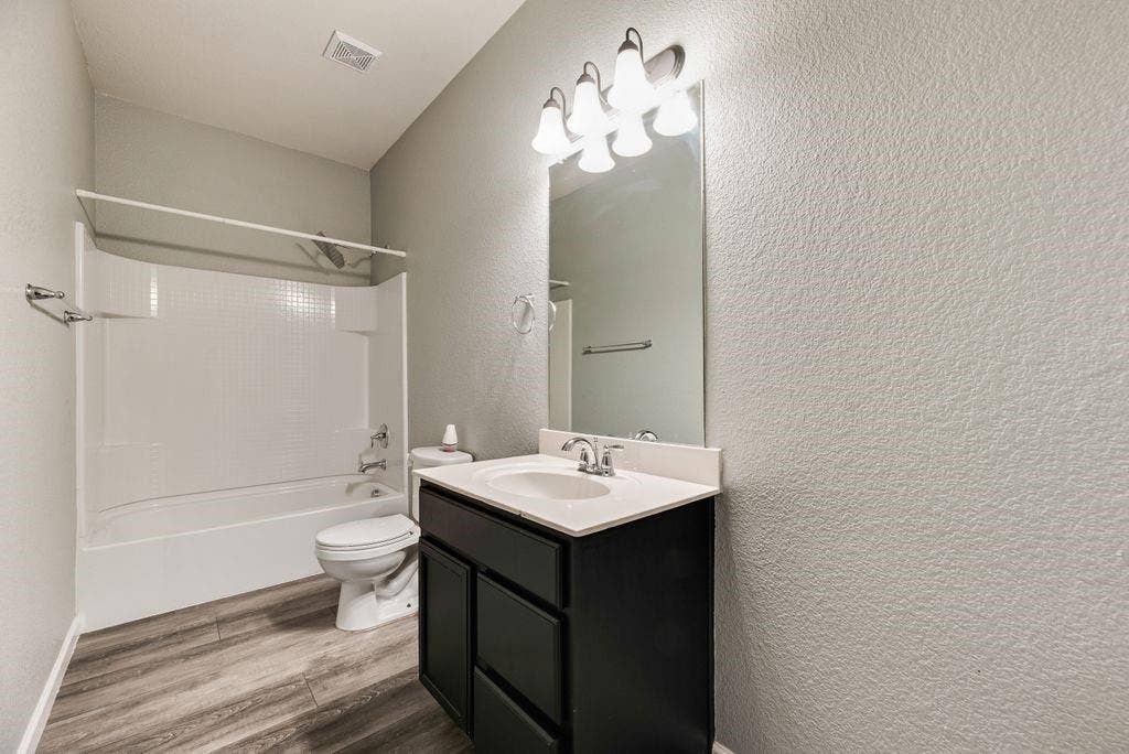 259 Saddlebrook Lane, Waxahachie, TX 75165 Modern bathroom featuring a black vanity with white rectangular sink and chrome fixtures, wood-finish flooring, large illuminated mirror with multi-bulb lighting, and a separate bathtub-shower combination with white subway tile surround and glass enclosure