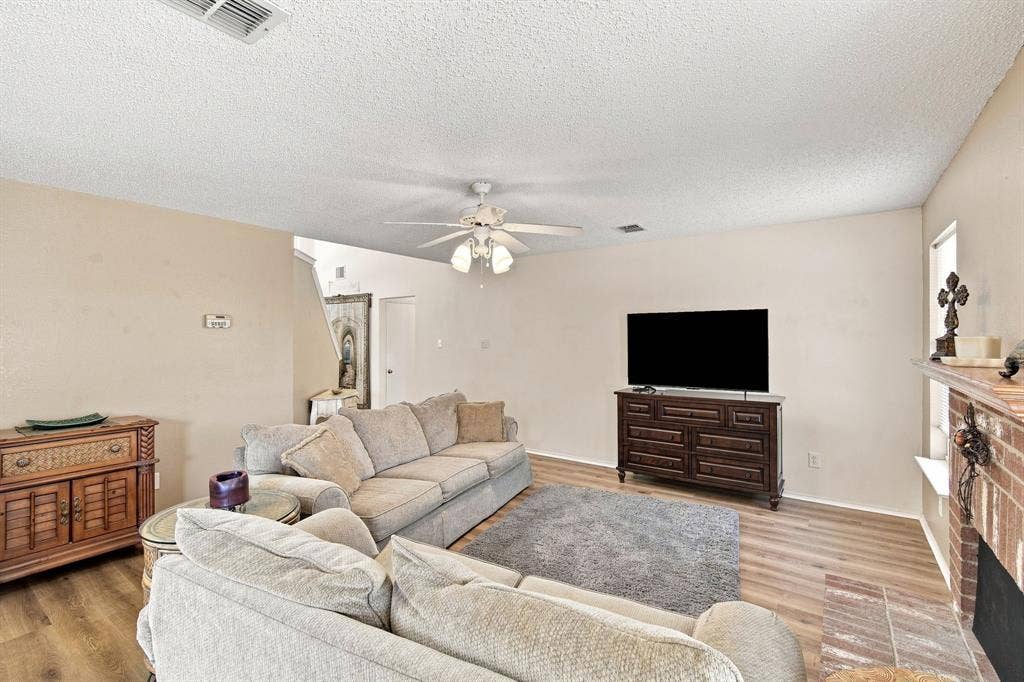 Wood-finish flooring, ceiling fan with integrated lighting, and a brick-faced fireplace with a mantel