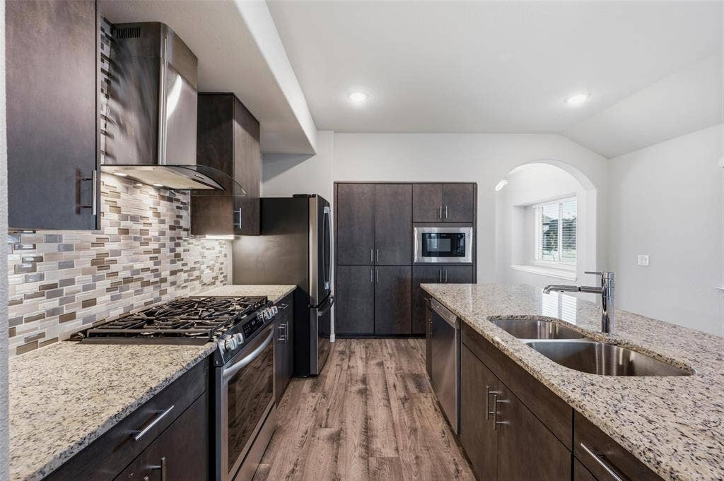 4425 Ridgehurst Lane, Fort Worth, TX 76036 Kitchen with wood-finish flooring, granite countertops, dark wood cabinetry, stainless steel appliances, and a mosaic tile backsplash