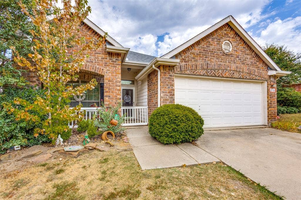2118 Hanakoa Falls Drive, Anna, TX 75409 Charming brick home featuring a covered front porch with white railings, two-car garage with white paneled door, manicured landscape with mature shrubs, concrete driveway, and gable roofline with architectural detailing