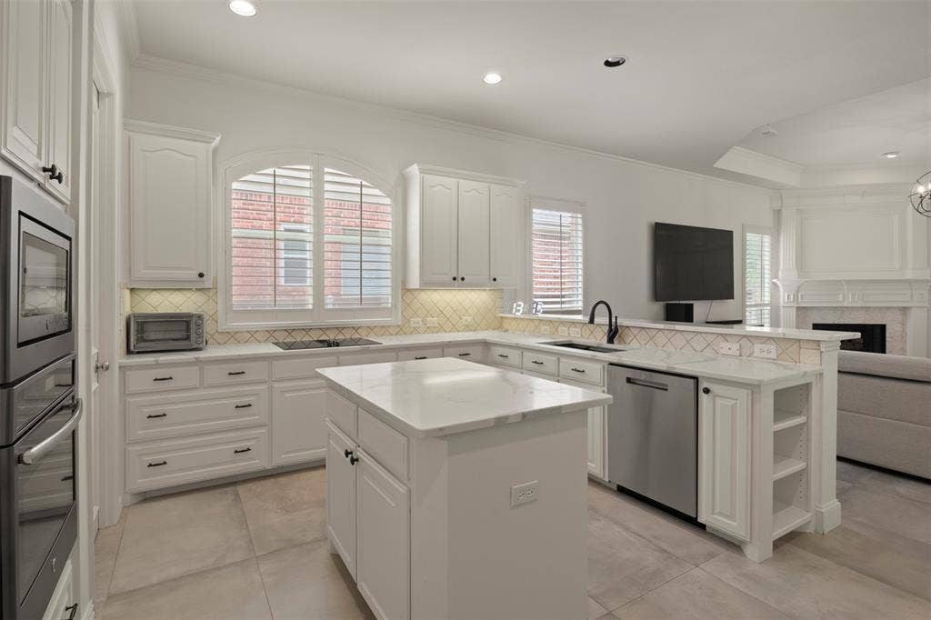 White cabinetry with dark hardware, a center island with a stone countertop, built-in double oven, integrated dishwasher, and a tile backsplash