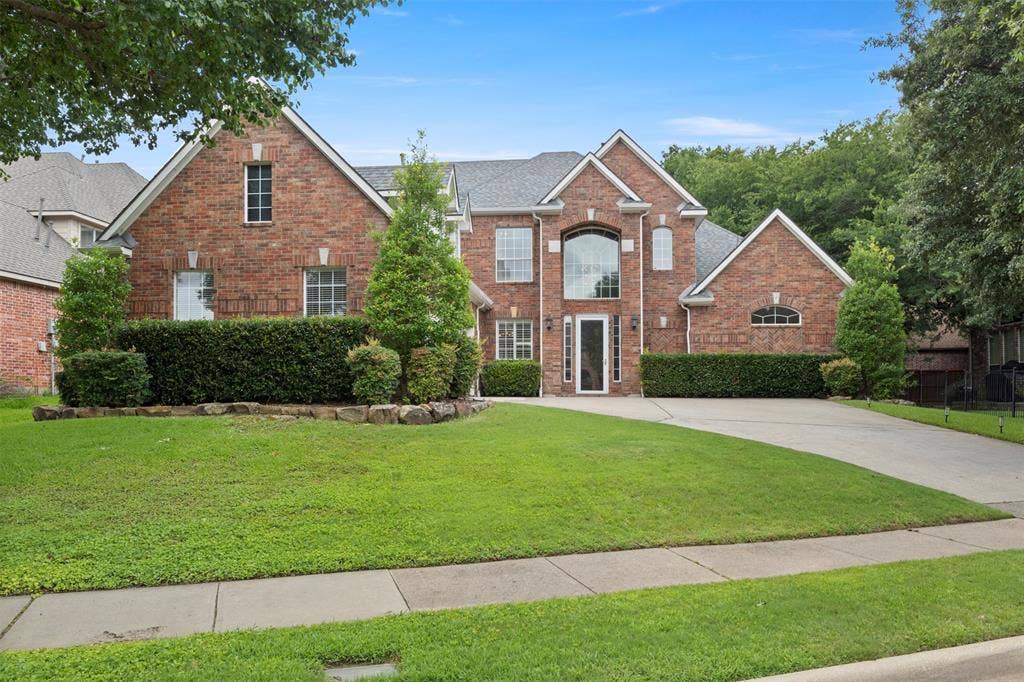 Brick exterior with a gabled roofline, arched window, mature landscaping, paved driveway, and established trees