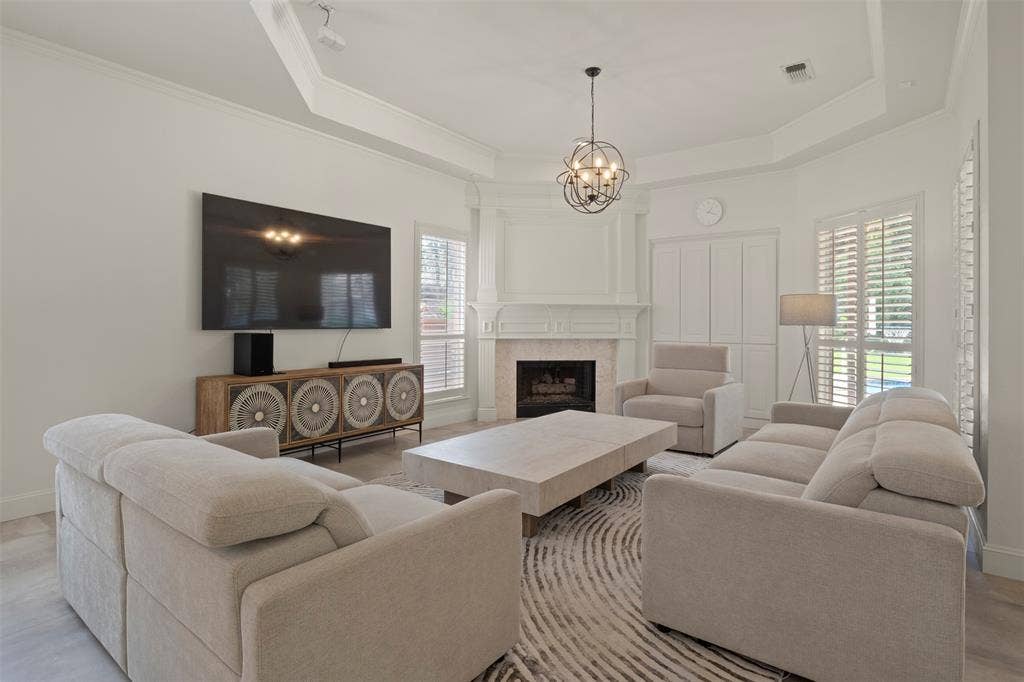 Tray ceiling with crown molding, corner fireplace with white mantel, plantation shutters, recessed lighting, and a contemporary chandelier