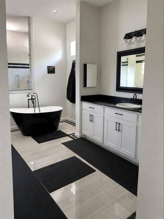 703 E Oates Road, Garland, TX 75043 Freestanding black soaking tub, dual-sink vanity with white cabinetry, dark countertops, and a framed mirror, and light-toned tile flooring with dark accents