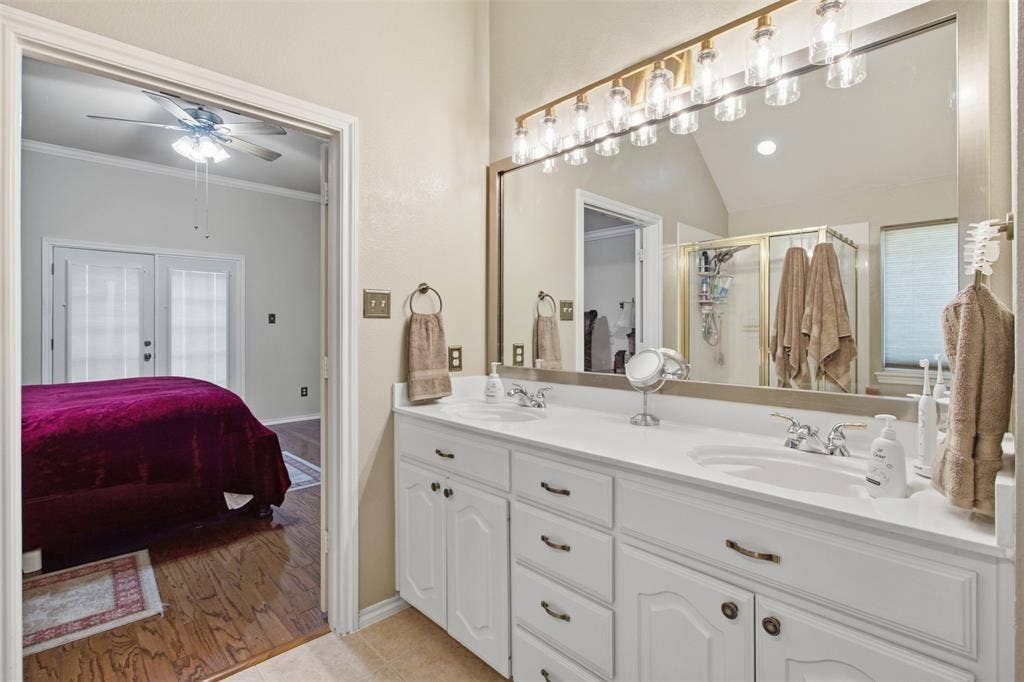 1112 Badbury Lane, Grand Prairie, TX 75052 Dual vanity with white cabinetry, polished chrome fixtures, and an expansive wall mirror with a multi-bulb light bar