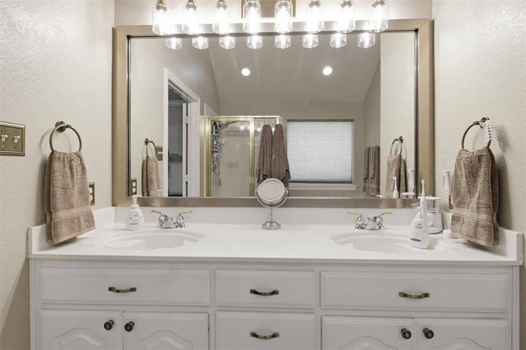 1112 Badbury Lane, Grand Prairie, TX 75052 Dual vanity with white cabinetry, integrated sinks, brushed nickel faucets, and a large framed mirror with overhead lighting
