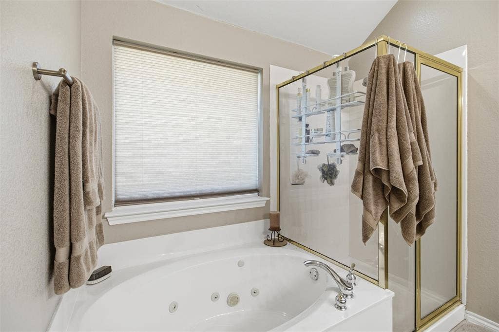 1112 Badbury Lane, Grand Prairie, TX 75052 Jetted tub with chrome fixtures, an adjacent glass-enclosed shower with a gold-tone frame, and a window with blinds