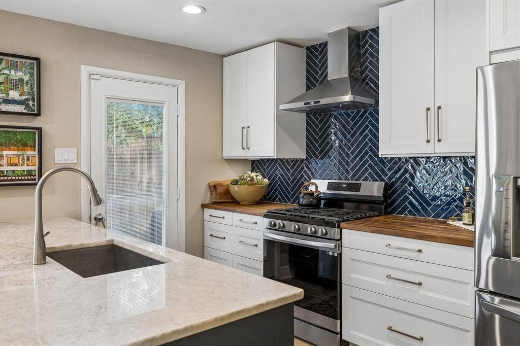 White shaker-style cabinetry with gold-tone hardware, wood-finish countertops, stainless steel appliances, herringbone-patterned tile backsplash, and a large single-basin sink