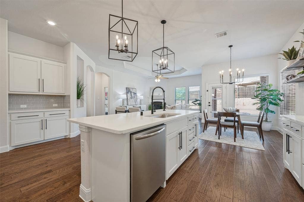 Kitchen featuring white cabinetry with dark hardware, a center island with a built-in sink and stainless steel dishwasher, wood-finish flooring, and a dining area with a modern chandelier