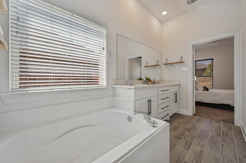 Soaking tub with a window, dual vanity with undermount sinks and quartz countertops, modern cabinetry with matte black hardware, and wood-finish flooring