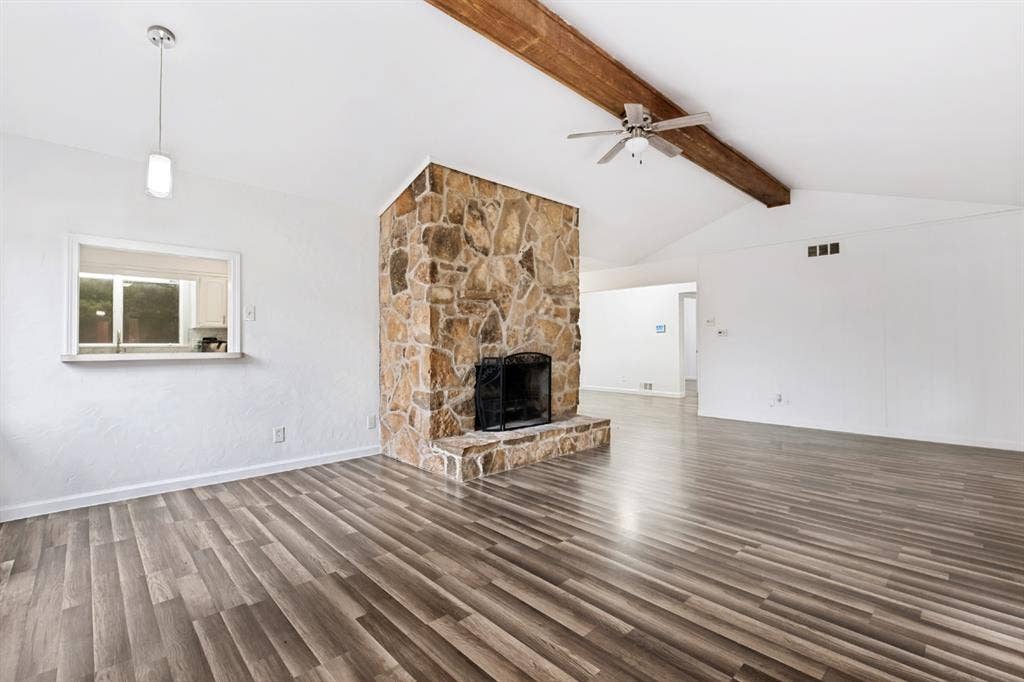 4315 Woodstock Drive, Dallas, TX 75232 Vaulted ceiling with exposed wood beam, wood-finish flooring, stone-clad fireplace with black grate, and a pass-through window