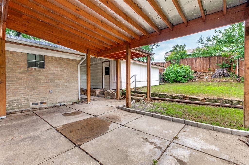 4315 Woodstock Drive, Dallas, TX 75232 Covered patio with exposed wood beam ceiling and brick exterior wall, featuring concrete flooring and direct access to a spacious fenced yard with mature landscaping and wooden fence perimeter