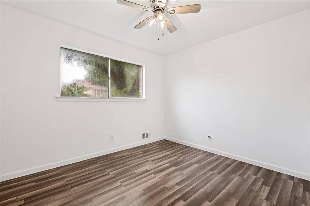 4315 Woodstock Drive, Dallas, TX 75232 Bright, light-filled bedroom showcasing newly installed wood-finish flooring, ceiling fan with integrated lighting, and large transom window providing natural illumination