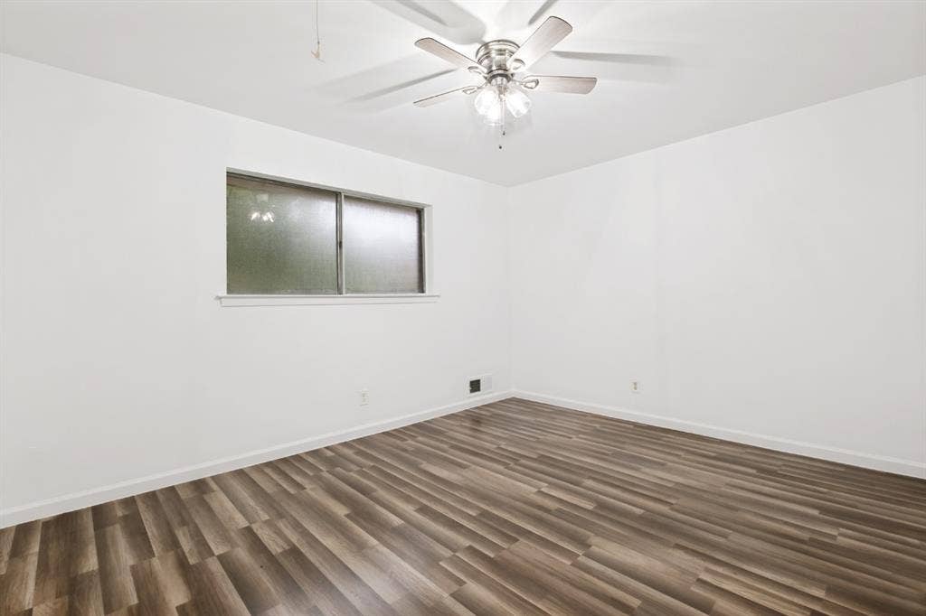 4315 Woodstock Drive, Dallas, TX 75232 Spacious bedroom showcasing wood-finish flooring with linear plank detailing, white walls, ceiling fan with integrated lighting, and a horizontal window with dual-pane glass providing natural light