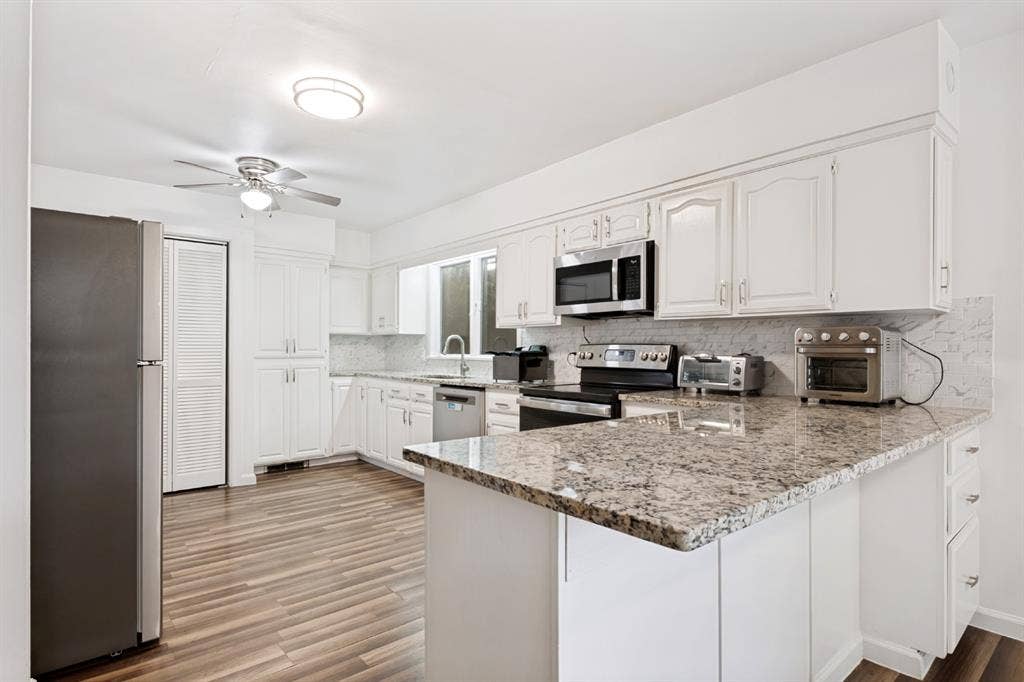 4315 Woodstock Drive, Dallas, TX 75232 Modern kitchen featuring white cabinetry with ample storage, granite-style countertops with waterfall island edge, stainless steel appliances including gas range and microwave, and wood-finish flooring throughout the open layout