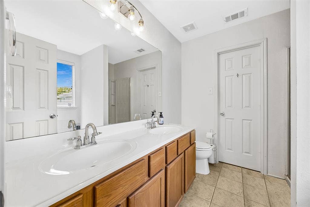Dual vanity with integrated sinks and chrome fixtures, wood-finish cabinetry, illuminated mirror, and tiled flooring