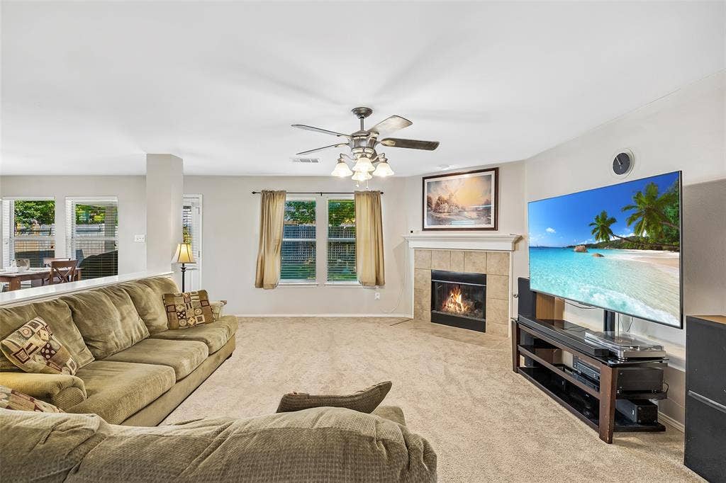 Carpeted living area featuring a tile-surround fireplace with mantel, ceiling fan with light fixture, and multiple windows