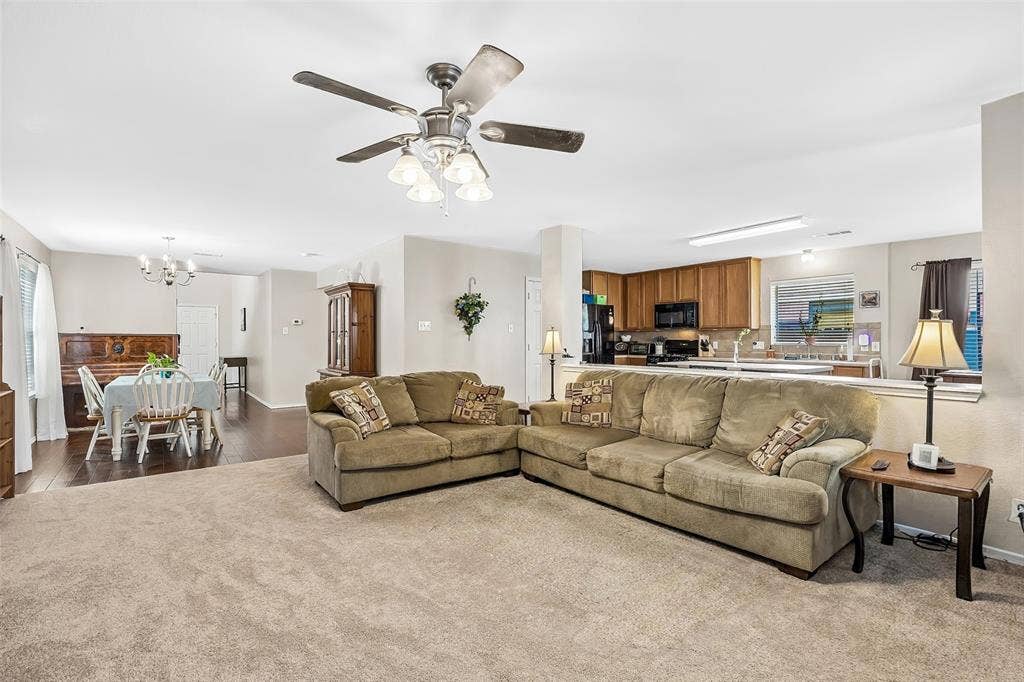 Expansive living area with neutral carpeting, an overhead ceiling fan with integrated lighting, and direct access to a kitchen featuring wood cabinetry and black appliances