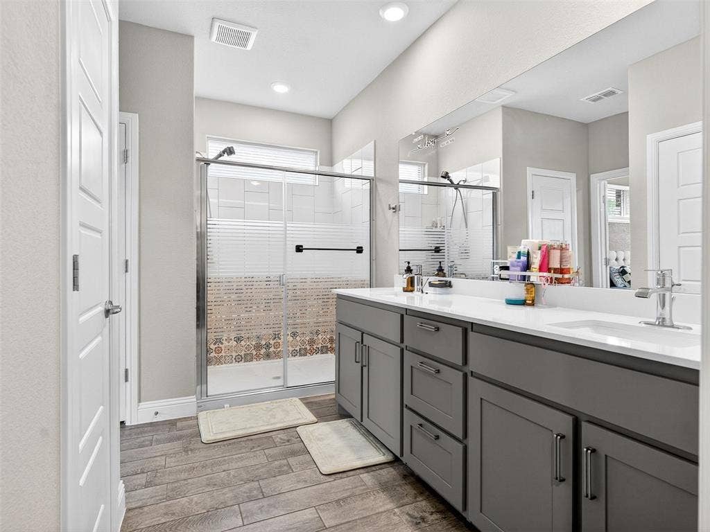 2504 Wren Lane, Denton, TX 76205 Dual vanity with under-sink storage, wood-finish flooring, and a glass-enclosed shower with decorative tiling