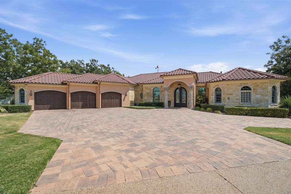Paver driveway, three-car garage with wood-finish doors, stone facade, arched windows, and a tiled roof