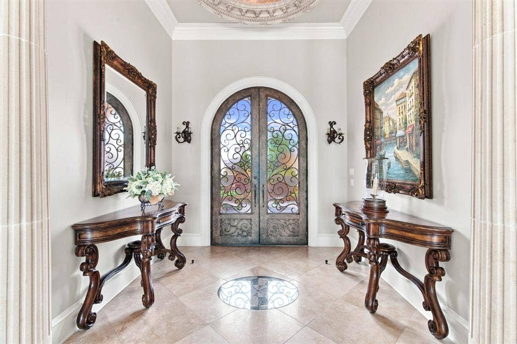 Arched entryway with ornate ironwork double doors, tile flooring with a decorative inlay, crown molding, and fluted architectural columns