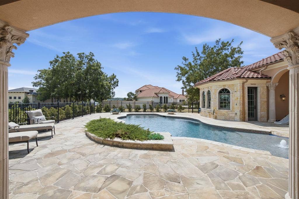Expansive flagstone patio with a curved swimming pool, surrounded by a wrought-iron fence and mature landscaping