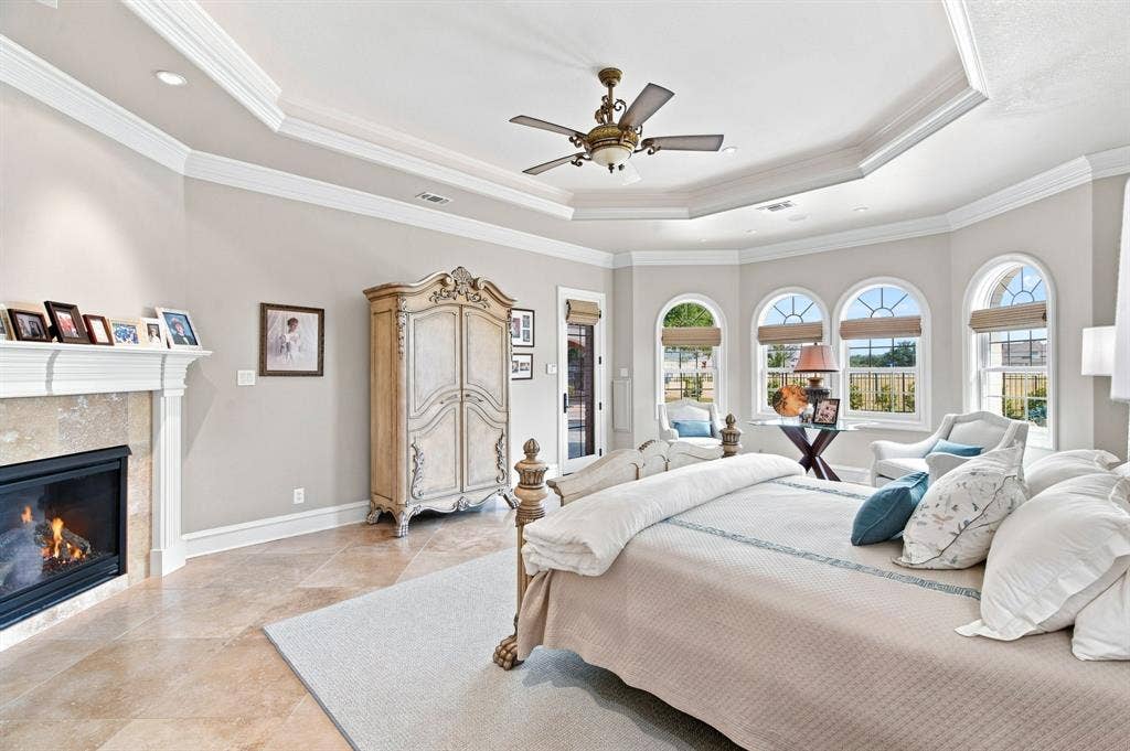 Tray ceiling with a decorative fan, a corner fireplace with stone surround, and light-toned tile flooring