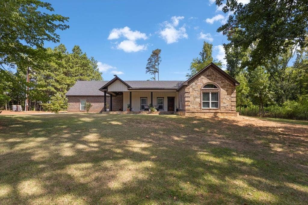 16136 County Road 1134=, Tyler, TX 75709 Stone and brick exterior residence with a covered porch, arched window, and dark shingle roof, set among mature trees