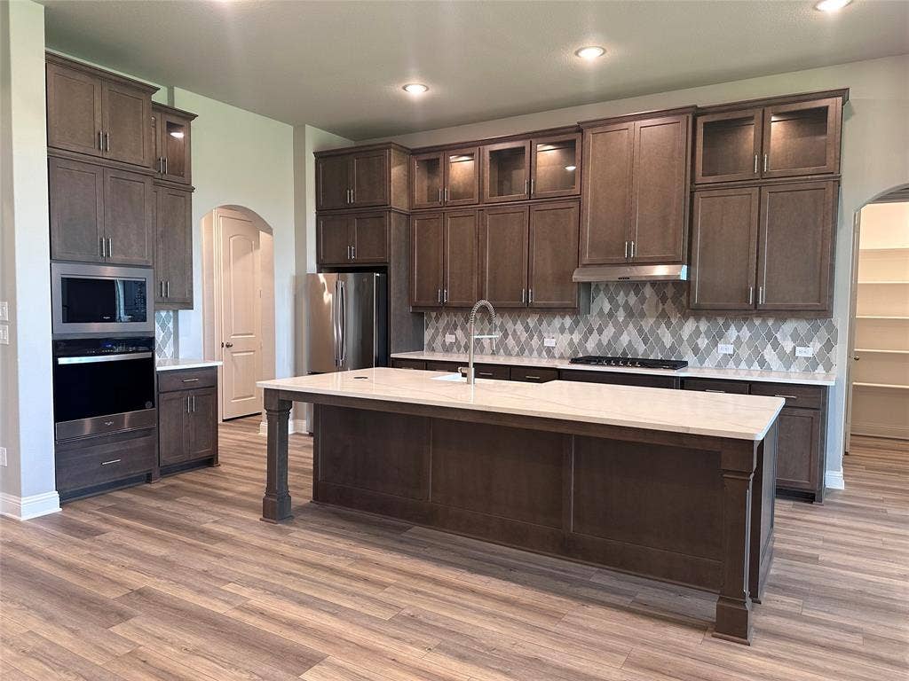2608 Roaming Trail, Northlake, TX 76247 Kitchen featuring a large central island with a light-toned countertop, extensive cabinetry with integrated lighting, stainless steel appliances, a geometric-patterned backsplash, and wood-finish flooring