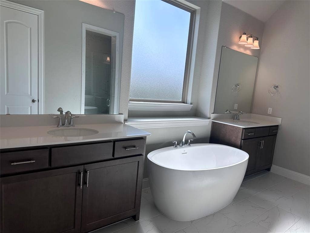 2608 Roaming Trail, Northlake, TX 76247 Dual vanities with white countertops, freestanding soaking tub, large frosted window, and light gray tiled flooring