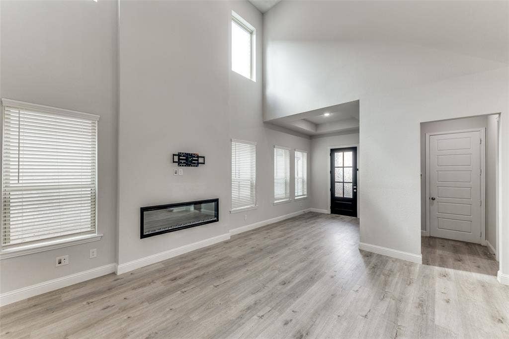 Unfurnished living room featuring a towering ceiling, plenty of natural light, a glass covered fireplace, light wood-style floors, and baseboards