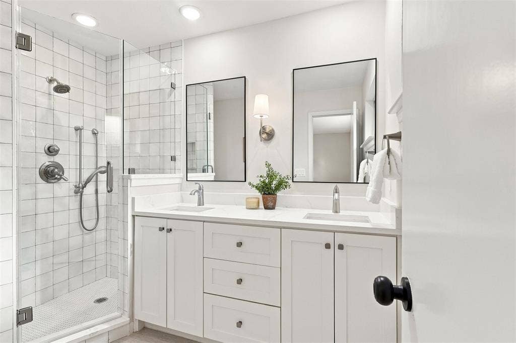 Glass-enclosed shower with subway tile surround, dual vanity with white cabinetry, quartz-finish countertops, and dual framed mirrors