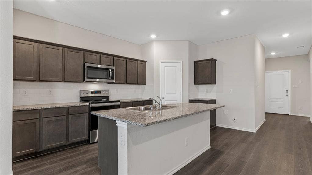 3037 Hidden Valley Drive, Lindale, TX 75771 Kitchen featuring dark wood-finish cabinetry, stainless steel appliances, a tile backsplash, and an island with a sink and stone countertop